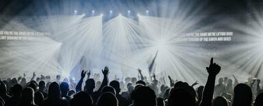 A crowd at the festival. Shapes of the attendees, many with their hands in the air, are standing out against the stage lights in the background